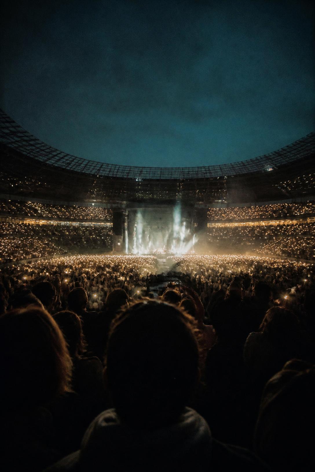A stadium concert at blue hour, tens of thousands of phone lights raised toward a single figure on a distant stage